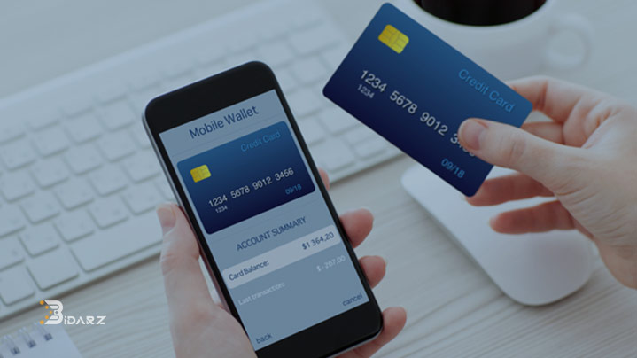 یک گوشی هوشمند با تصویر یک کارت اعتباری در کنار دست یک انسان که کارتی مشابه را در دست دارد. صفحه کلید و کاوس سفید رنگ در پشت تصویر پیدا هستند.